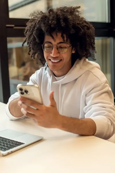 Student met bril en krullen zit aan tafel en kijkt lachend op zijn telefoon.
