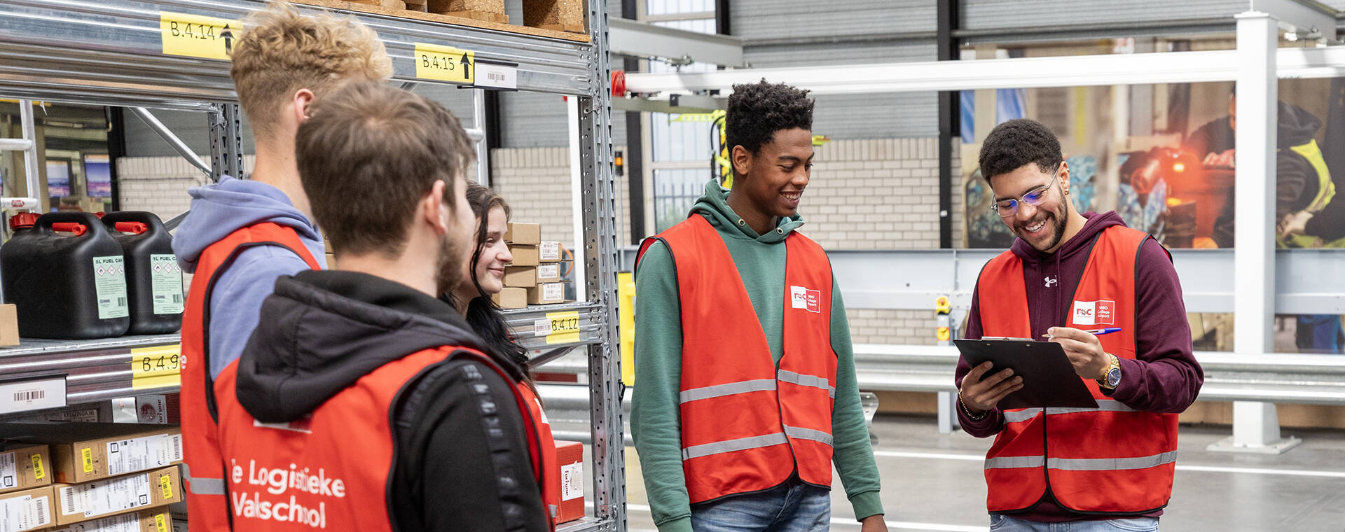 Meerdere studenten in rode hesjes staan in een opslagloods bij elkaar.