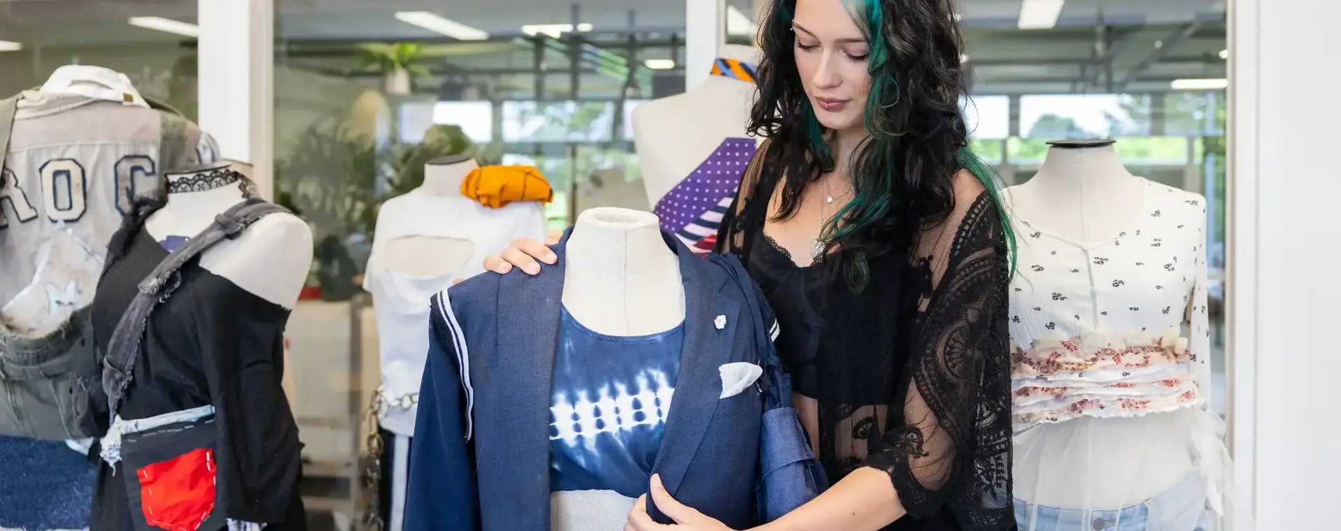 Student modeontwerp bekijkt een blauwe outfit op een paspop in het atelier. Op de achtergrond staan meer paspoppen met kledingstukken.