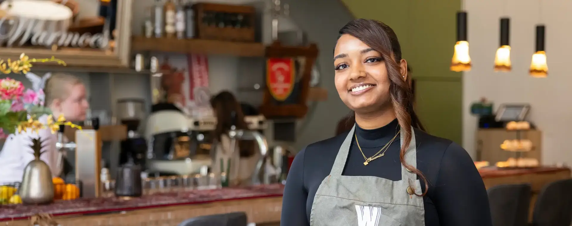 Vrouwelijke horecamedewerker met schort staat glimlachend in een restaurant.