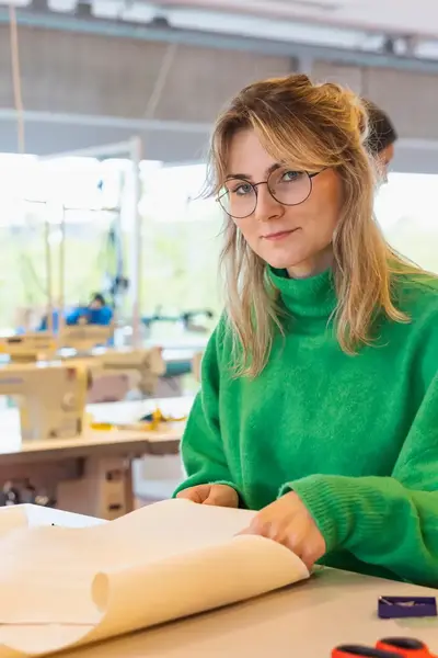 Jonge vrouw met groen trui aan achter tekentafel met op de achtergrond naaimachines.