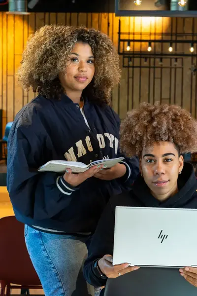 Twee studenten werken samen aan een opdracht in een gezellige leeromgeving. Eén student houdt een laptop vast, terwijl de ander aantekeningen maakt in een notitieboek.