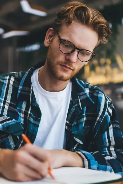 Student met bril en geruit overhemd schrijft geconcentreerd in een notitieboek.