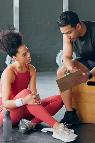 Sportinstructeur bekijkt samen met een vrouwelijke sporter een schema. Ze zitten in een gym of fintessruimte.