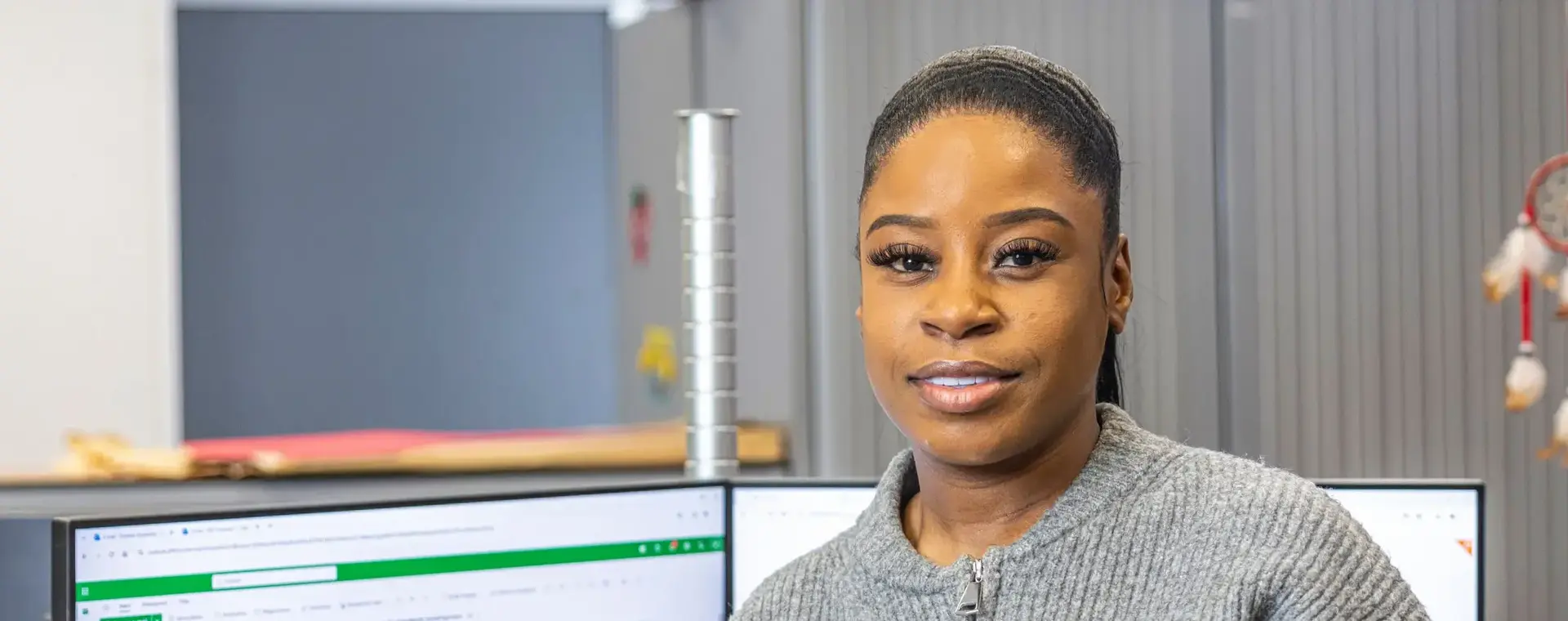 Een vrouw met een headset in haar hand leunt tegen haar bureau. Op haar bureau staan twee computerschermen die aan staan. 