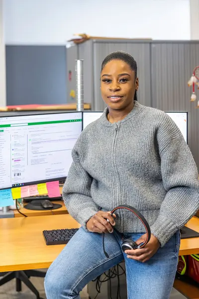 Een vrouw met een headset in haar hand leunt tegen haar bureau. Op haar bureau staan twee computerschermen die aan staan. 
