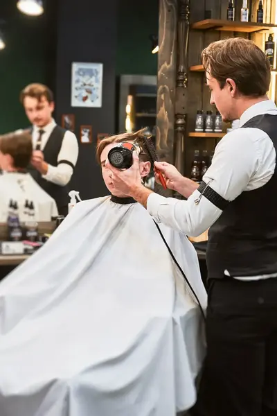 Klant in kappersstoel wordt geföhnd door kapper in kapsalon. 