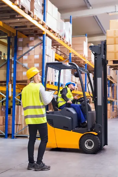 Twee collega's in gele hesjes halen order op met vorkheftruck in magazijn. 