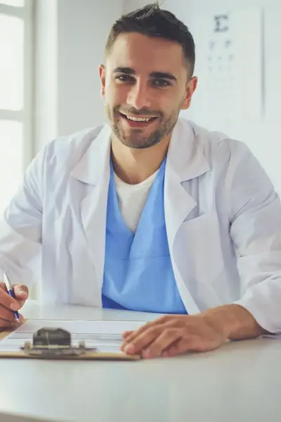 Dokter zit glimlachend achter bureau met laptop en notitieblok.