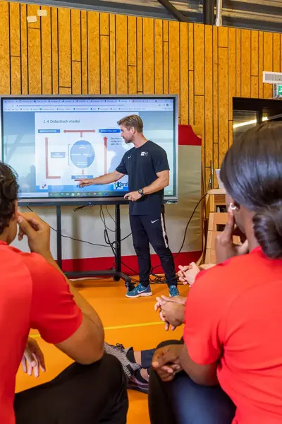 Groep studenten in rode shirts met 'Sport & Bewegen' luistert in de gymzaal naar een docent die bij een scherm uitleg geeft met een presentatie.
