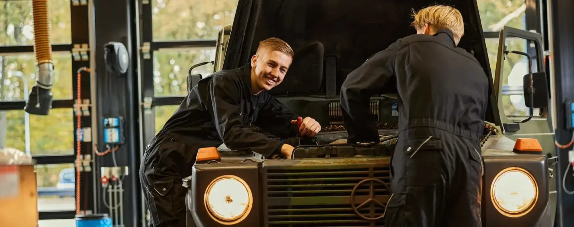 Twee studenten sleutelen aan een legervoertuig in een werkplaats, een van hen kijkt lachend naar de camera.
