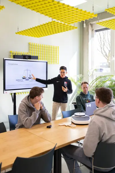 Student geeft uitleg aan studenten in een klaslokaal en wijst naar een presentatie op een scherm.