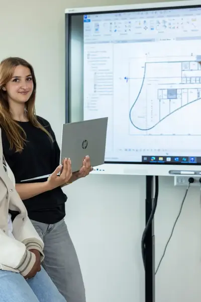 Twee studenten in klaslokaal met laptop in de hand voor een scherm met daarop een bouwtekening.
