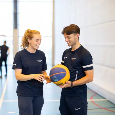 Twee studenten in sportkleding praten en lachen samen in de gymzaal terwijl ze een basketbal vasthouden.