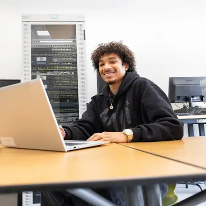 Student werkt op een laptop in een ICT-lokaal met netwerkapparatuur op de achtergrond. 