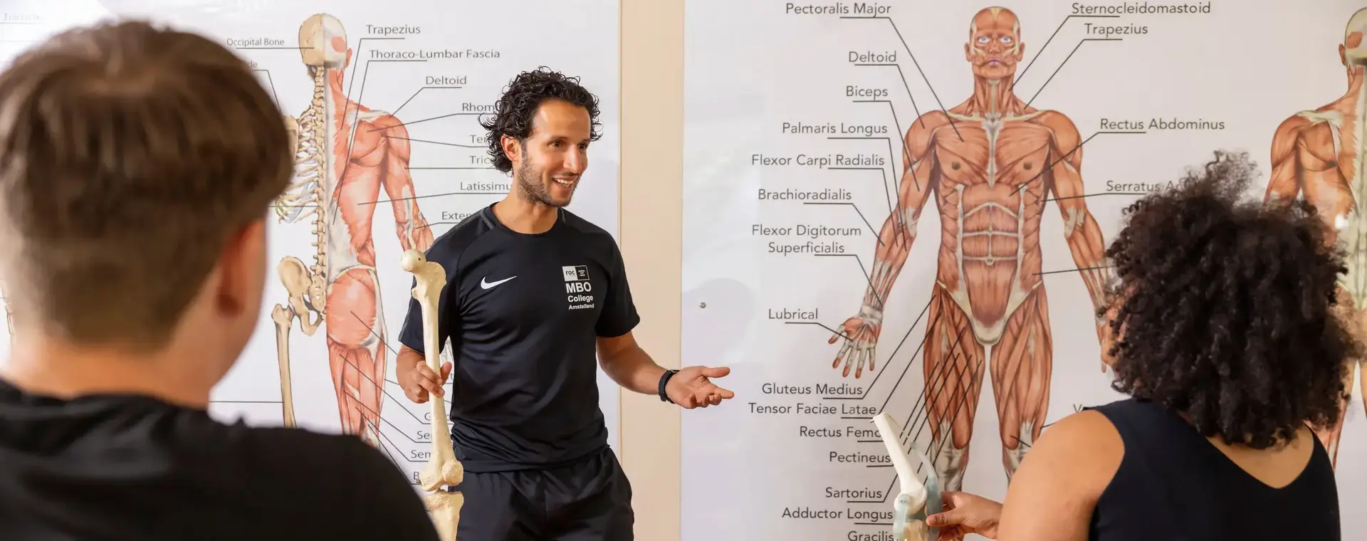Sportdocent toont deel geraamte aan klas met anatomietekeningen op de achtergrond