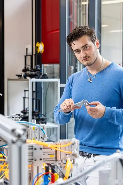 Een jongeman in een blauwe trui staat geconcentreerd bij een technisch werkstation en houdt een schuifmaat vast. Hij is bezig met het nauwkeurig meten van een onderdeel in een techniekomgeving.