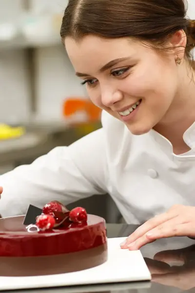 Student decoreert een rode taart met chocolade en kersen. Ze werkt nauwkeurig in de bakkerijkeuken.
