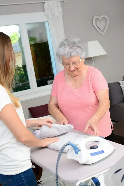 Student helpt een oudere vrouw met strijken in de woonkamer.