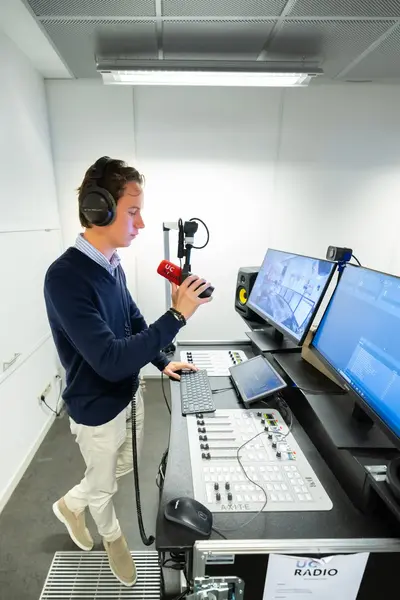 Student met een koptelefoon op staat in een studio en praat in een microfoon terwijl hij naar meerdere beeldschermen kijkt.