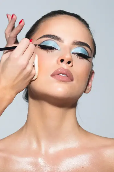 Student visagie brengt nauwkeurig blauwe oogschaduw en eyeliner aan bij een model tijdens een praktijkles.