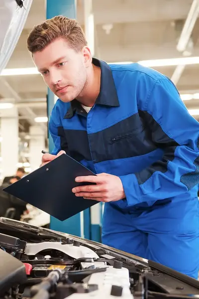Automonteur in overall controleert auto onder de motorkap met clipboard